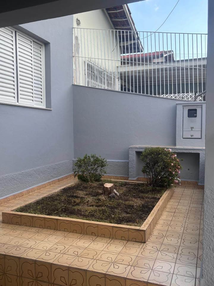Área externa com piso estampado, pequeno canteiro central com vegetação, paredes cinzas e muros altos com grades.