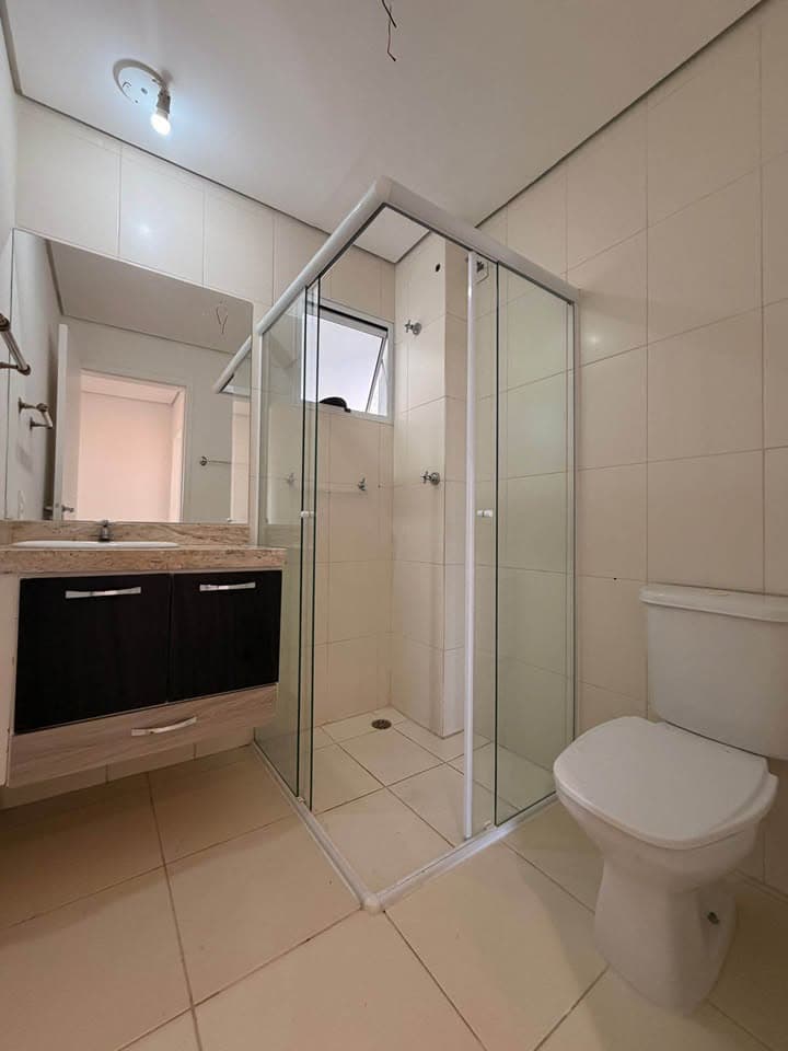 Banheiro com revestimento em cerâmica clara, box de vidro, vaso sanitário e pia com gabinete escuro.