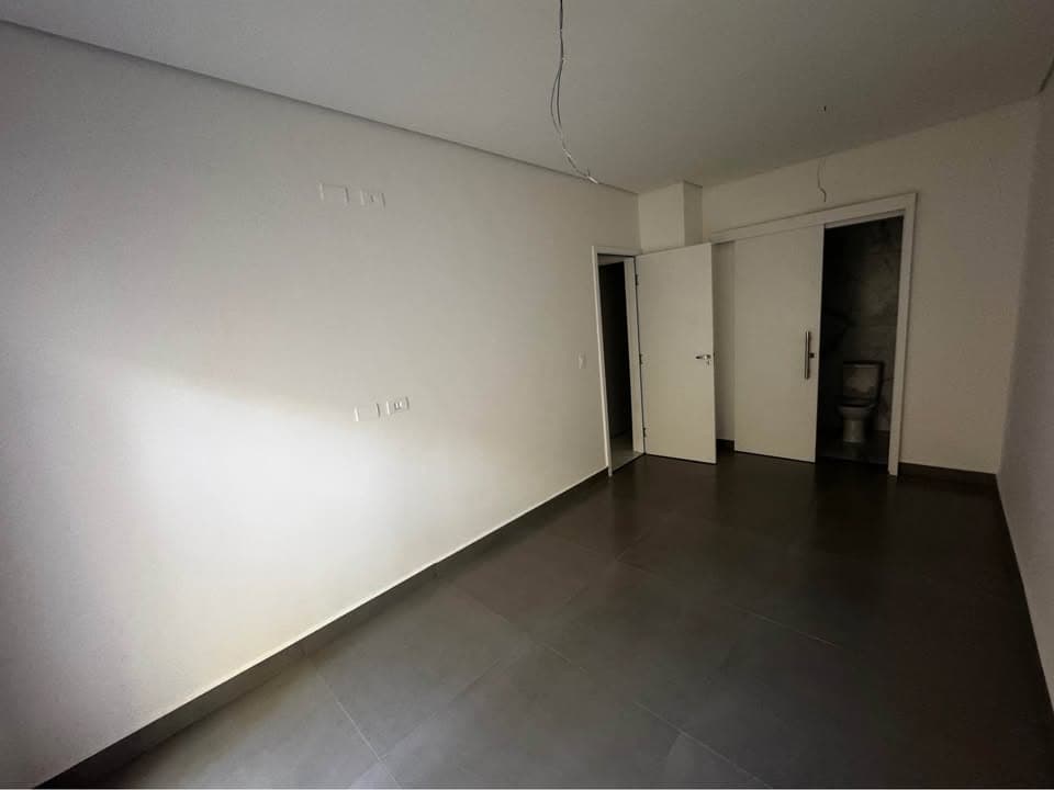 Quarto com piso escuro, paredes brancas e porta de correr dando acesso ao banheiro.